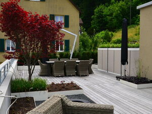 . Eine Terrasse mit Holzboden, geflochtenen Stühlen, Tisch, Pflanzen, rotem Baum und einem beige Gebäude im Hintergrund.