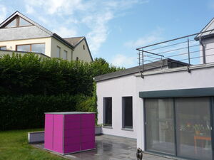 . The image shows a modern house with a flat roof and a balcony with metal railing. There is a brightly colored structure, possibly a storage unit or decorative element, in magenta on the patio. The house has large windows and is surrounded by greenery. Another house with a pitched roof is visible in the background. The sky is mostly clear with a few clouds.