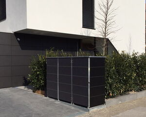 . The image shows a modern building exterior with a black wall and a black structure that seems to be a utility box or cabinet. There is a hedge, young trees, and a paved area in front. There's also a shadow from a lamppost on the pavement.