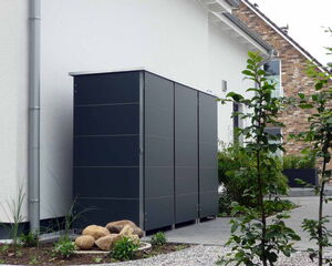 . The image shows an outdoor setting with a modern storage cabinet or shed next to a building. The cabinet is made of dark panels. There are plants and rocks nearby, and the building appears to have a brick façade. The area is landscaped with gravel and pathways.