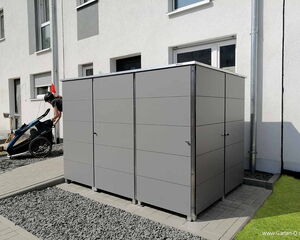 . The image shows a modern storage unit or shed placed outdoors on a paved area with gravel and grass surrounding it. The structure appears to have metal or composite paneling with doors featuring handles. In the background, there is a person wearing a helmet, possibly with a child trailer bike. The walls of a building and a rainwater downpipe are also visible.