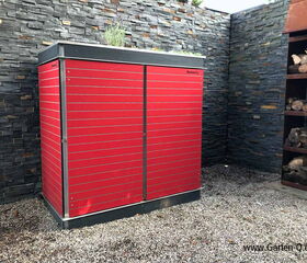 . The image features a red storage structure known as "Garten Q." It appears to be placed outdoors against a wall made of stacked stones. To the right, there's a wood rack containing cut logs. The ground is covered with gravel. The structure seems to be designed for garden or outdoor use.