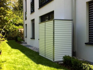 . The image shows the side of a building with a neat, well-maintained lawn in the foreground. There are several windows with closed blinds on the side of the building. A large, rectangular, horizontal-slatted metal structure is attached to the wall, likely used for covering air conditioning units or storage. Surrounding the area are bushes and trees, indicating a landscaped garden.