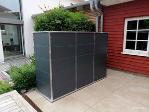 . Eine moderne, rechteckige Gartenbox mit Pflanzen oben, auf einer Terrasse neben einem roten Gebäude.