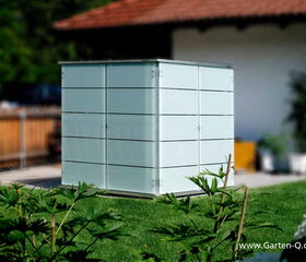 . The image features a sleek, modern storage unit situated outdoors on a lawn. The structure is cube-shaped with a minimalist design, possibly used for storing garden tools or equipment. It appears to be made of metal or a similar material, and it's set against a backdrop of a garden with some plants and a building nearby.