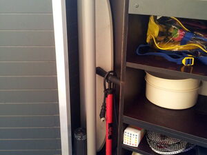 . The image shows a storage cabinet containing the following items:

1. An ironing board.
2. A bicycle pump.
3. Various tools and objects on shelves, including:
   - A box or container.
   - A package with a multicolored cube pattern (possibly a calendar or note pad).
   - A yellow bag with items inside.
   - A white bowl or pot.
4. A wheeled base or stand at the bottom.

The cabinet has several shelves organizing the items.