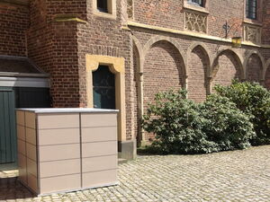 . The image depicts a tall, rectangular storage unit placed in front of a historical brick building with arched windows. The building has ornate detailing, and there is greenery, such as bushes, alongside the stone-paved ground.