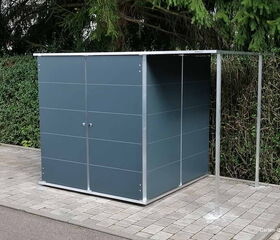. Ein moderner Gartenschuppen aus Metall auf einem gepflasterten Untergrund, umgeben von Bäumen und Büschen.