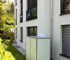 . The image shows an outdoor storage unit situated beside a modern building. The building has several large windows with blinds, and the unit is positioned on a well-maintained lawn. The scene includes a neatly arranged garden area with greenery and plants. The website "www.Garten-Q.de" is visible on the bottom right of the image.