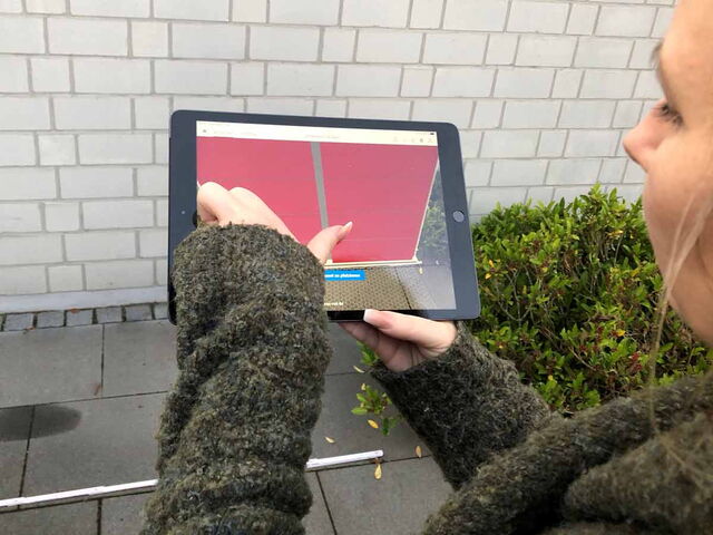 Eine Person hält ein Tablet und betrachtet ein Bild mit einem roten Objekt darauf. Im Hintergrund eine gemauerte Wand und Pflanzen.