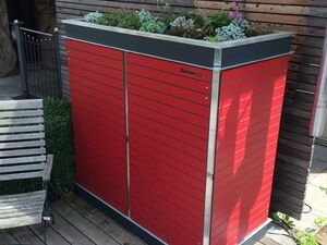 . Roter Gartenschrank mit Pflanzen auf dem Dach, auf einer Holzterrasse neben einem Holzzaun und einer Bank.