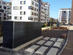 . The image shows a modern residential area with several apartment buildings surrounded by landscaped areas. In the foreground, there is a structure, possibly a shelter or storage unit, made of dark panels. In front of it are rectangular stepping stones placed on the ground. The background includes other buildings of varying heights, a truck, and some construction materials, indicating ongoing development or landscaping work. The overall setting appears clean and urban.