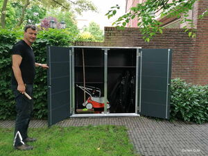 . Ein Mann öffnet einen Gartenschrank mit Rasenmäher, Besen und gefalteten Stühlen.