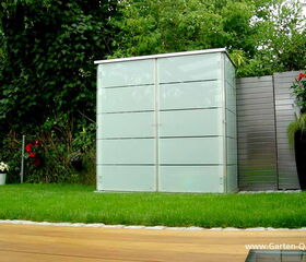 . The image shows a garden scene with a modern outdoor storage unit. The storage unit is rectangular with a sleek, minimalist design featuring horizontal panels. It is surrounded by greenery, flowers in pots, and a wooden deck in the foreground. The website "www.Garten-Q.de" is visible in the bottom right corner.