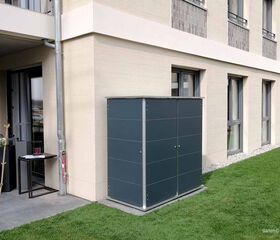 . Gebäudefassade mit Fenstern, graues Metallschuppen im Garten, Tisch mit Papieren daneben.