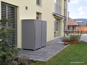 . Ein grauer Gartenschrank steht auf einer Terrasse neben einem modernen Haus mit Garten.