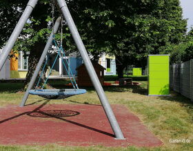 Ein Spielplatz mit Nestschaukel, Schuppen, grünem Schrank, Rasenflächen und Bäumen im Hintergrund.