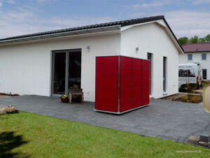 . The image shows a modern outdoor setting with a single-story house featuring a tiled patio. On the patio, there is a large red storage unit or cabinet with multiple panels. The house has a sleek design with white walls and dark-framed windows. In the foreground, there is a grassy lawn, and in the background, there are more houses and a vehicle with signage. There are some potted plants and outdoor decor near the house.