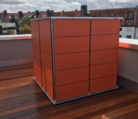 . Ein orangefarbener Kasten auf einer Dachterrasse mit Holzboden und Wolken am Himmel.