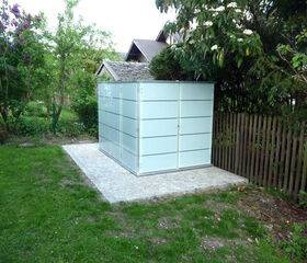 . The image shows a small, modern glass structure with frosted panels, possibly a shed or storage unit, situated in a garden. It is surrounded by grass, trees, flowers, and a wooden fence. There's also a paved area where the structure is placed.
