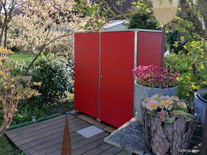 . Ein roter Gartenschrank im Grünen, umgeben von Blumen in Töpfen und Holzböden darunter.