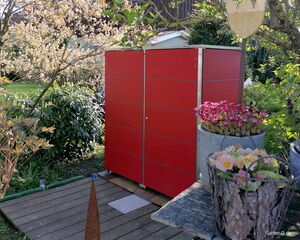 Rotes Gartenhaus im Grünen, umgeben von Pflanzen und Blumenarrangements.