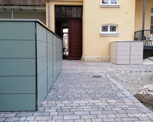 Hinterhof mit gelbem Gebäude, grauen Schränken und gepflastertem Weg.