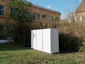 . Ein weißer Gartenschrank steht auf einer Wiese vor einem Backsteingebäude, umgeben von Büschen und Bäumen.