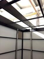 The image shows the interior of a structure with metal framework and white panel walls. The ceiling has exposed beams and there are cables or wires visible at the top. The setting appears industrial or construction-like.
