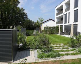 . The image shows a modern residential outdoor space featuring a multi-story white building with balconies. In front of the building is a well-maintained garden area with neatly trimmed grass, shrubs, and small trees. There are paved pathways and several dark-colored outdoor storage units or sheds. The setting looks calm and organized, with patio seating areas that include sun umbrellas and furniture for outdoor relaxation.