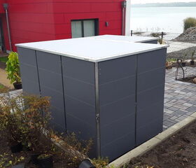 . The image shows an outdoor scene with a modern building featuring a red wall and black and gray elements. In the foreground, there's a cubic structure with a gray exterior, possibly a garden storage unit or shed. The area around it includes a paved walkway, plants, and landscaping work in progress. Nearby, there's a body of water and a person working on the landscaping.