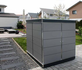 . Ein moderner grauer Gartenschrank steht in einem gepflegten Garten mit Terrasse, Häusern und Kiesweg.