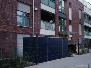 . The image shows a modern building with a brick facade, multiple windows, and external roller shutters. In the foreground, there is a structure with dark panels and a metal fence surrounding it. It looks like a storage unit or enclosure next to the walkway leading to the building. Vegetation is visible along the fence.