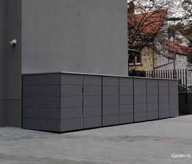 Graue Schränke stehen an einer Außenwand, umgeben von Gehwegplatten und Bäumen im Hintergrund.