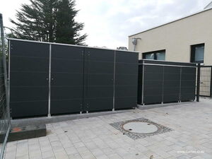 . Schwarze Mülltonnenboxen vor einem Gebäude auf einem gepflasterten Hof.