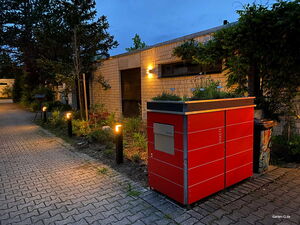 . Ein beleuchteter Weg mit rotem Gartencontainer, Pflanzen darauf, daneben eine Mauer mit Fenstern und Pflanzen.