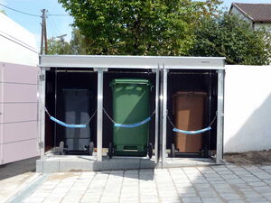 . The image shows an outdoor enclosure containing three different colored waste bins. Each bin has its own compartment within a metal structure, and they are separated by dividers. The structure appears to keep the bins organized and secure, possibly for recycling purposes.