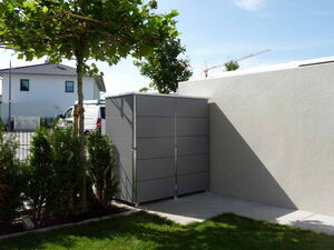 . Ein grauer Schuppen neben einer weißen Wand im Garten, mit Bäumen und Gras umgeben.