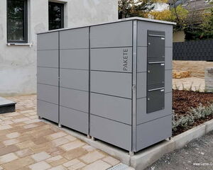 . The image shows a modern outdoor parcel locker system. It has a sleek, gray design with multiple compartments for receiving packages. It's located in an outdoor setting next to a building.