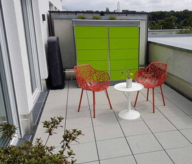 . The image shows a small outdoor patio area. It includes two red chairs with intricate, openwork designs and a small white round table between them. On the table, there is a small vase with a few flowers. In the background, there is a bright green cabinet or storage unit. The patio is paved with light gray tiles, and there are plants visible in the foreground and background.