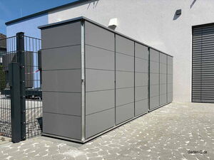 . Eine moderne, graue Mülltonnenbox mit Lamellenstruktur neben einem Gebäude und Zaun.