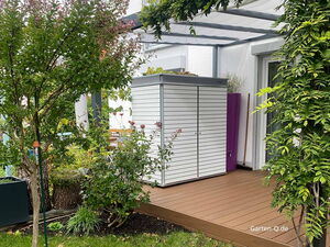 Gartenschrank mit einer Dachbegrünung auf einer Terrasse, grünem Rasen, Pflanzen und einem weißen Schuppen unter einer Pergola.