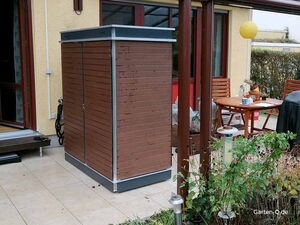 . Ein Gartenhaus aus Holz steht auf einer Terrasse neben einem Tisch mit Stühlen und Pflanzen.