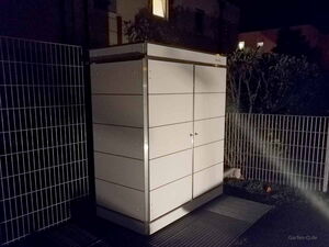 . Ein Gartenhaus oder Geräteschuppen aus Metall, beleuchtet in der Nacht, umgeben von einem Gitterzaun.