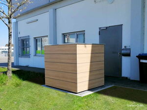 . Holzverkleidete Box vor einem Gebäude mit Tür, Fenstern und Mülltonne auf einer Wiese.
