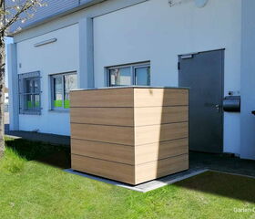 . Holzverkleidete Box vor einem Gebäude mit Tür, Fenstern und Mülltonne auf einer Wiese.