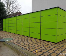 Grüner Garten-Schuppen auf gepflasterter Fläche neben einem Gebäude, Baum im Hintergrund.