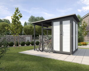 The image depicts a modern garden shed with a bicycle storage area on a small paved section. The shed has white panels with a dark frame and a flat roof. Several bicycles are parked outside under an overhang. The surrounding area includes neatly trimmed grass, trees, and a brick wall in the background. There is a house visible to the right.
