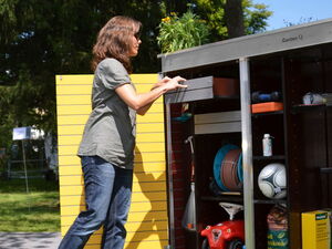 . The image shows a person interacting with a storage shed or cabinet labeled "Garten Q." The cabinet is outdoor, likely used for garden or yard equipment. Visible items include a ball, a toy vehicle, a broom, a hose reel, and various garden tools and supplies. There are plants on top of the cabinet, and the person appears to be accessing or organizing items within it.
