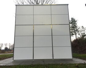 . The image shows a structure that resembles a minimalist, modern cube-like building or installation. The surface is divided into panels, and it is outdoors, surrounded by grass and some trees. It appears to be made of light-colored materials, possibly metal or composite panels.